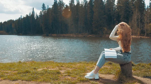 Bella giovane donna bionda seduta a leggere un libro in un parco vicino al lago. Vista laterale — Foto Stock