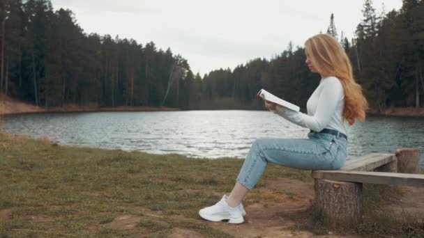 Schöne junge blonde Frau sitzt und liest Buch in einem Park in der Nähe des Sees. Seitenansicht — Stockvideo