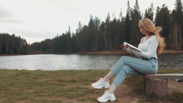 Hermosa joven rubia sentada y leyendo libro en un parque cerca del lago. Vista lateral — Vídeo de stock