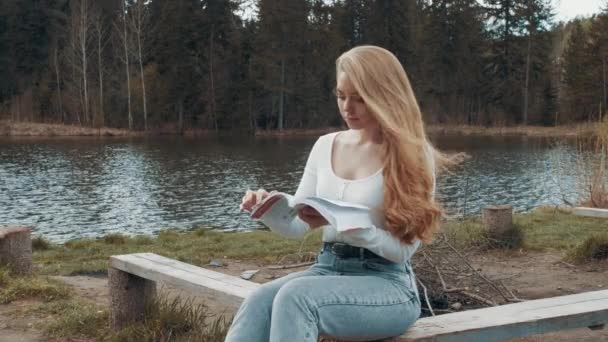 Bella giovane donna bionda seduta a leggere un libro in un parco vicino al lago. Vista laterale — Video Stock