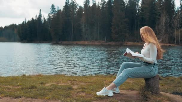 아름다운 금발의 젊은 여자가 호수 근처 공원에 앉아서 책을 읽고 있습니다. 옆면 — 비디오