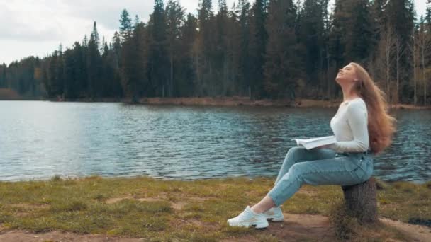 Hermosa joven rubia sentada y leyendo libro en un parque cerca del lago. Vista lateral — Vídeos de Stock