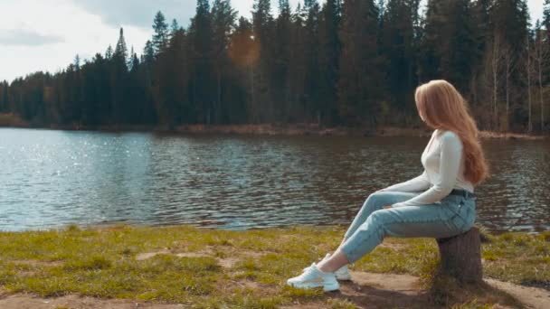 Hermosa joven rubia sentada y relajarse en un parque cerca del lago. Vista lateral — Vídeo de stock