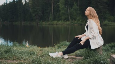 Güzel sarışın kadın, doğal görünüşü, uzun saçları, güzel yüzü, gölün yanındaki parkta oturuyor. Yan görünüm