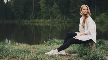 Güzel sarışın kadın, doğal görünüşü, uzun saçları, güzel yüzü, gölün yanındaki parkta oturuyor. Yan görünüm