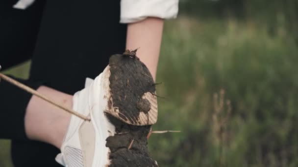 Девушка в белой рубашке и белых кроссовках очищает грязь от подошвы стоя на одной ноге, закрывая — стоковое видео
