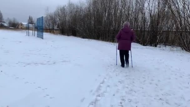 高齢の女性は学校のスタジアムの雪道を歩くノルディックのための棒で歩く — ストック動画