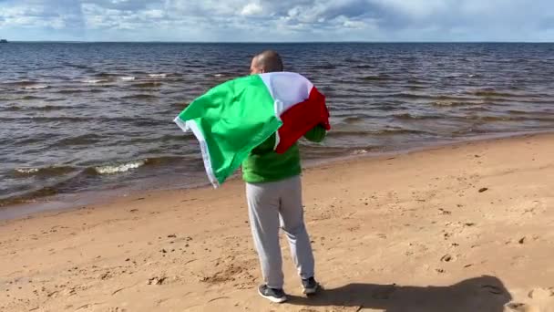後ろにイタリア国旗を手にしたハンサムな男 晴れた日に一人のイタリア人が海沿いに散歩 — ストック動画