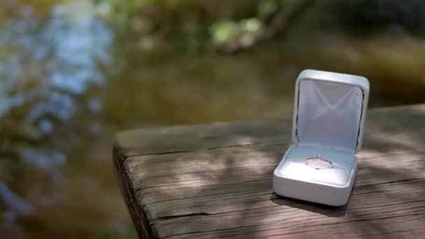 Sortija de compromiso de oro en caja blanca se sienta en una mesa de madera junto al río, empuja la muñeca — Vídeos de Stock