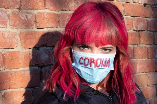 Girl with mask to protect her from Corona virus. Corona virus pandemic. Corona written on mask. Woman with mask standing next to brick wall. Beautiful red haired girl with medical mask.