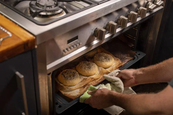 Chef reaching with two hands to pull freshly baked bagels out of a hot oven