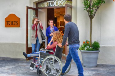 İSPAN, LLORET DE MAR - 14 Nisan 2019: Village Outlet 'in arka planında kırık bacaklı bir kızı sandalyeye taşıyan bir adam