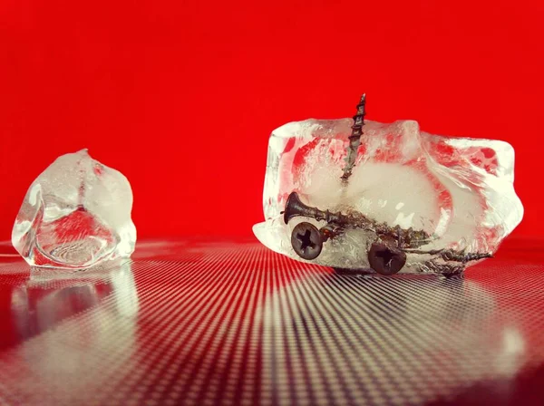 A cold shard of ice with metal screws frozen in it on a textured surface and a red background in a creative macro photo