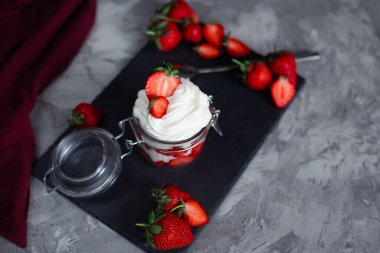 Çilek ve krema tatlısı şeffaf cam bir kavanozda çimento ve siyah levha tezgahında, büyük çileklerle süslenmiş.