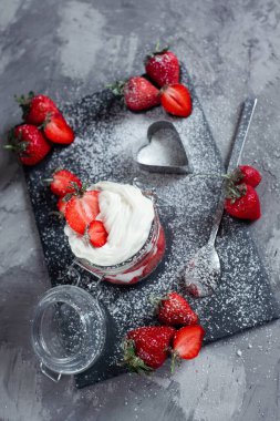 Çilek ve krema tatlısı şeffaf cam bir kavanozda çimento ve siyah bir levha tezgahında, büyük çileklerle süslenmiş ve pudra şekeriyle serpilmiş.