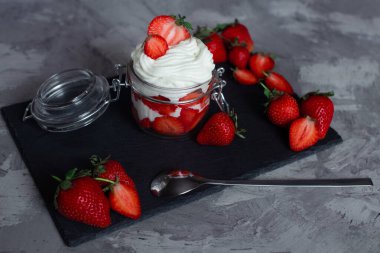 Çilek ve krema tatlısı şeffaf cam bir kavanozda çimento ve siyah levha tezgahında, büyük çileklerle süslenmiş.
