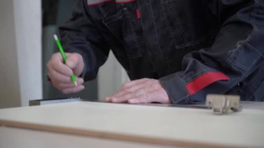 İş kıyafetleri içinde çalışan yetişkin bir erkek işçi, büyük bir marangoz masasında mobilya yapmak için çizim yapıyor. Bir adamın yüzünün yakın çekimi..