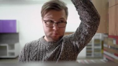 Sakallı ve gözlüklü bir adam, kazak giyiyor, mutfakta duruyor ve rafı açıyor ve içine bakıyor..