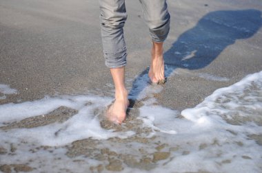 Bir adam kumsalın altın kumu boyunca yürüyor. Denize yakın yürüyen erkek bacakları. Kumlu bir sahilde dalgalarla yürüyen çıplak ayaklı bir adam. Yaz tatili ya da tatil.