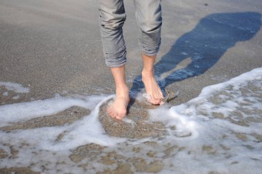 Bir adam kumsalın altın kumu boyunca yürüyor. Denize yakın yürüyen erkek bacakları. Kumlu bir sahilde dalgalarla yürüyen çıplak ayaklı bir adam. Yaz tatili ya da tatil.