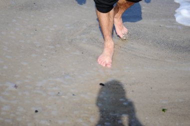 Bir adam kumsalın altın kumu boyunca yürüyor. Denize yakın yürüyen erkek bacakları. Kumlu bir sahilde dalgalarla yürüyen çıplak ayaklı bir adam. Yaz tatili ya da tatil.