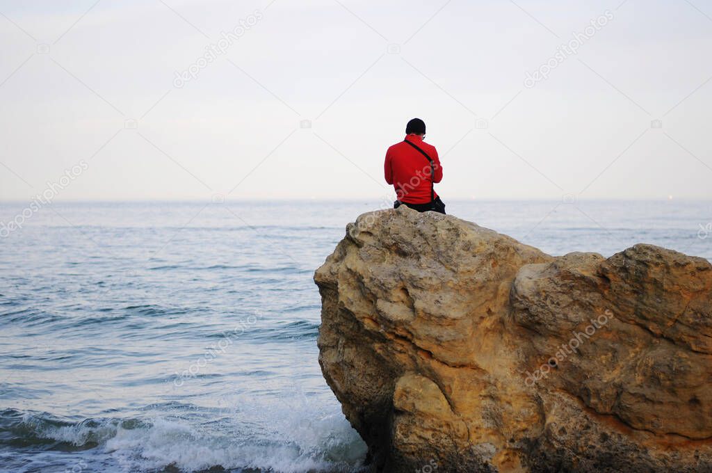One guy sits on a rock by the sea and looking sunset.