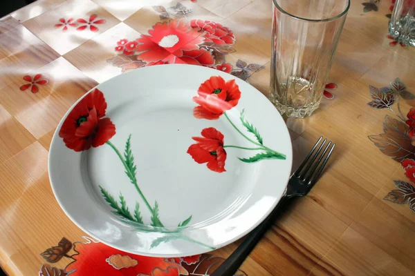 Kırmızı haşhaş boyalı beyaz tabak ve masanın üzerinde cam bardak..