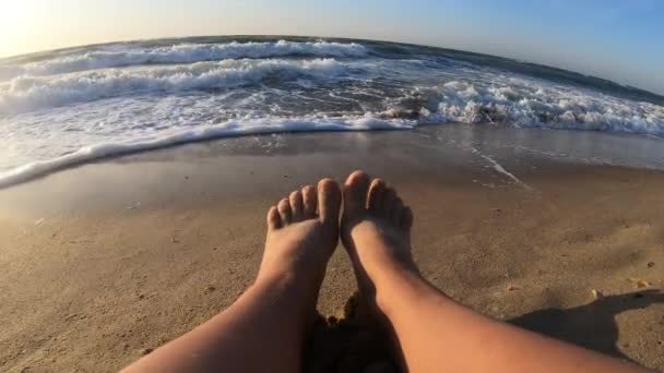 해변에 앉아 있는 소녀는 바다 근처에 앉아 있고 파도가 모래 위에서 발을 구르고 있다 — 비디오