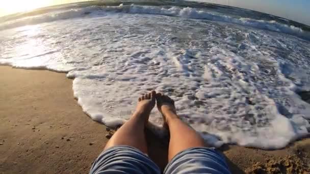 A menina está sentada perto do mar e ondas rolam em seus pés na praia arenosa — Vídeo de Stock