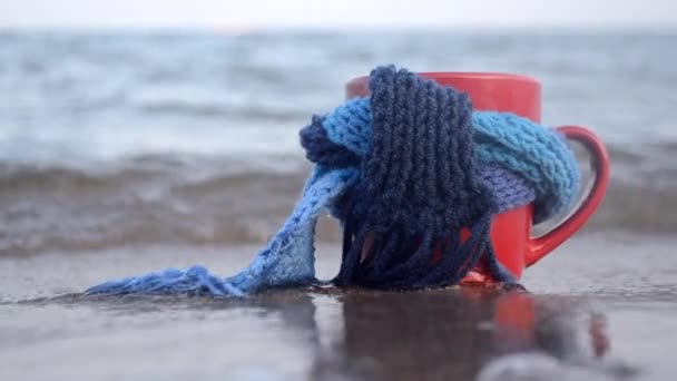 Červený hrnek s kávou svázaný s modrým pleteným šátkem stojí na písečné pláži — Stock video