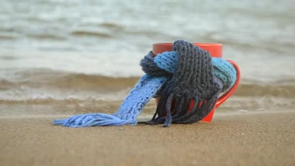 Red mug with coffee tied with blue knitted scarf stands on sandy beach — Stock Video