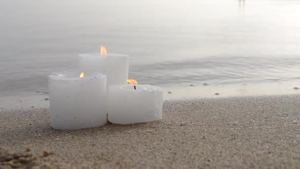 Three small white paraffin candles burning on sandy beach shore edge — ストック動画