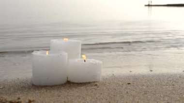 Three small white paraffin candles burning on sandy beach shore edge