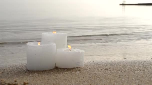 Three small white paraffin candles burning on sandy beach shore edge — Wideo stockowe