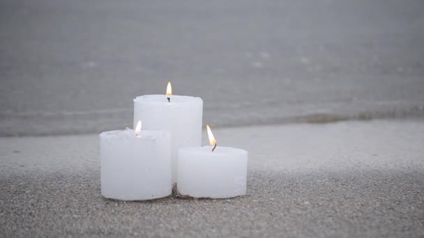 Three small white paraffin candles burning on sandy beach shore edge — 图库视频影像