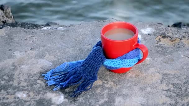 Червоний кухоль з кавою і парою пов'язаний з синім в'язаним шарфом стоїть на камені — стокове відео