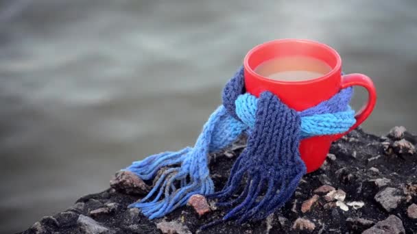 Red mug with coffee and steam tied with blue knitted scarf stands on stone — Stockvideo