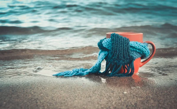 Red mug with coffee tied with blue knitted scarf stands on sandy beach — Stock Photo, Image