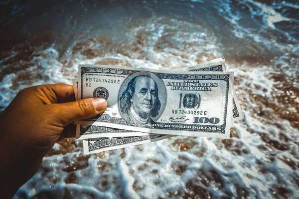 Girl holding money bill of 300 dollars on background of sea oceans waves and sand wet beach close-up on sunny day. Hand waves sea ocean money dollars vacation. Concept finance money holiday traveling