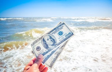 Güneşli bir günde deniz dalgaları ve gökyüzünün arka planında 300 dolarlık para tutan bir kız. El dalgaları okyanus para dolar tatil tatil dinlenme yaz. Finansal para tatil ufkunu kavramsallaştır