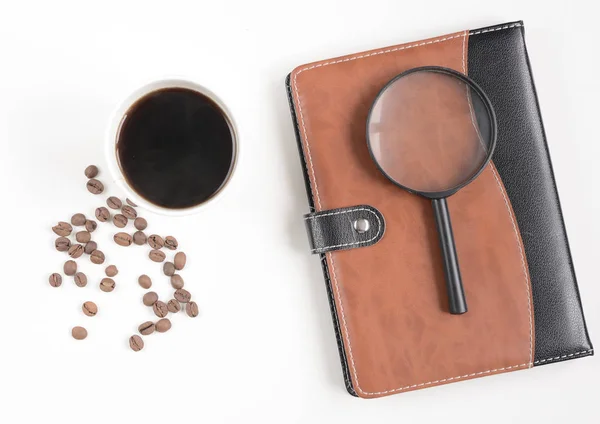 Xícara de café e grãos de café com notebook e lupa n mesa branca — Fotografia de Stock