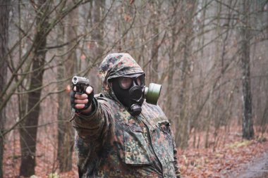 Kamuflajlı bir asker ve ormanda elinde bir silah, gaz maskesi takıyor.