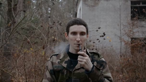 在秋天的森林里 一个年轻的士兵在一座废弃的建筑物的背景下抽烟斗的画像 — 图库视频影像