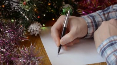 Bir adam Noel ağacının yanındaki masaya ellerini uzatır ve Noel Baba 'ya yeni yıl mektubu yazar.