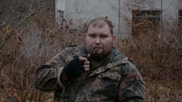 Retrato de um homem cheio em roupas de camuflagem, um silvicultor que fuma um cachimbo de tabaco — Vídeo de Stock