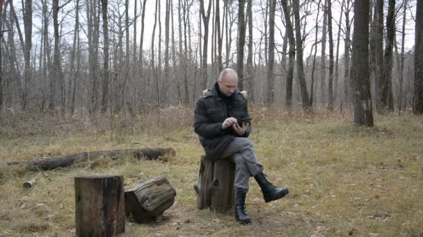 Un viajero se sienta en un bosque en un tocón y escribe sms en una tableta — Vídeos de Stock
