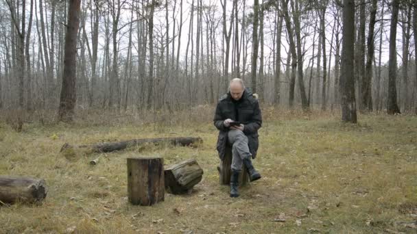 Een man zit in het herfstbos en typt tekst op een tablet — Stockvideo