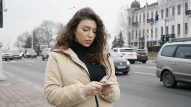 Kıvırcık saçlı güzel esmer kız işlek bir şehir yolunun yakınındaki akıllı telefonuna bir mesaj yazıyor.