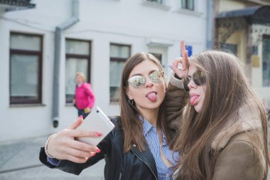 İki güzel genç kadın fotoğraf çekerken eğleniyor ve cep telefonuyla yüzünü buruşturuyor, yaşam tarzı kavramı.
