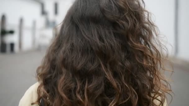 Une fille aux cheveux bruns avec de longs cheveux bouclés marche dans la rue en regardant avec un dos au ralenti — Video